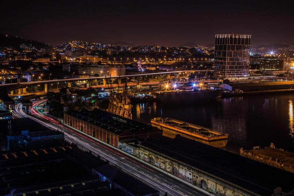 Vista aérea nocturna de un puerto urbano, mostrando estelas de luz de larga exposición de coches en una autopista junto a barcos atracados.
