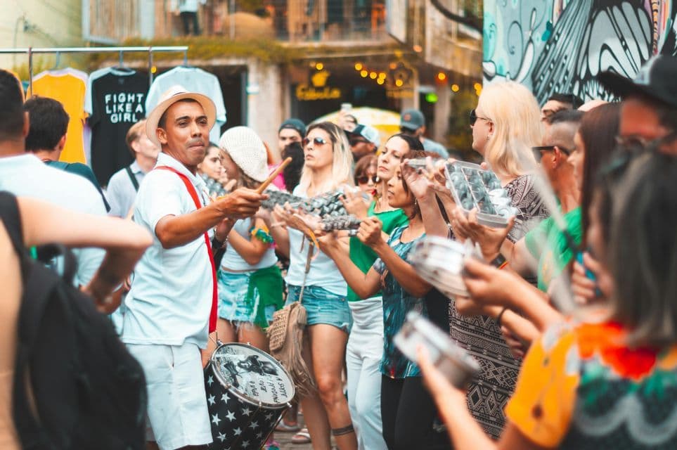 Un músico con sombrero lidera a una multitud de personas tocando instrumentos de percusión durante un festival callejero.
