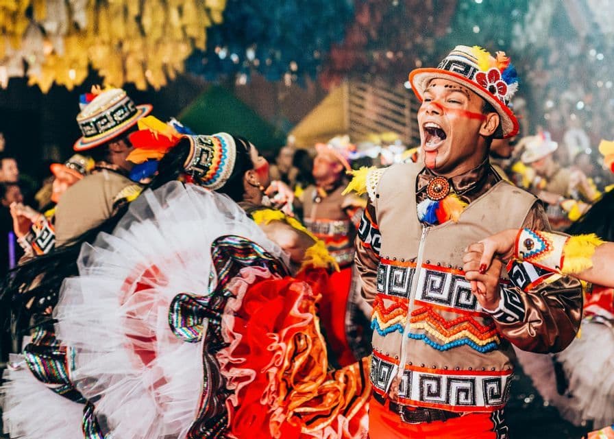 Un hombre con un sombrero de plumas colorido y un chaleco de cuentas canta mientras baila con otras personas durante un festival.