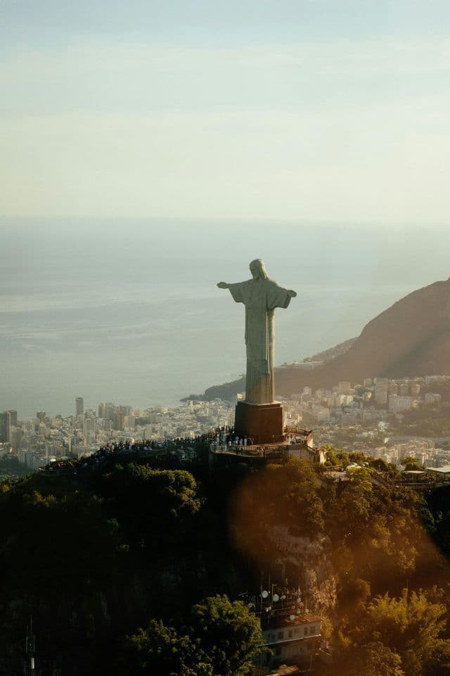 Una gran estatua con los brazos extendidos se alza en la cima de una montaña verde, dominando una ciudad costera con una multitud de gente en su base.