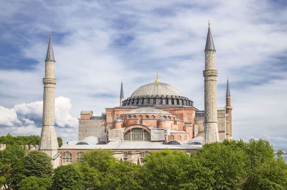 A large mosque with a central dome and four minarets stands above green trees against a partly cloudy sky.