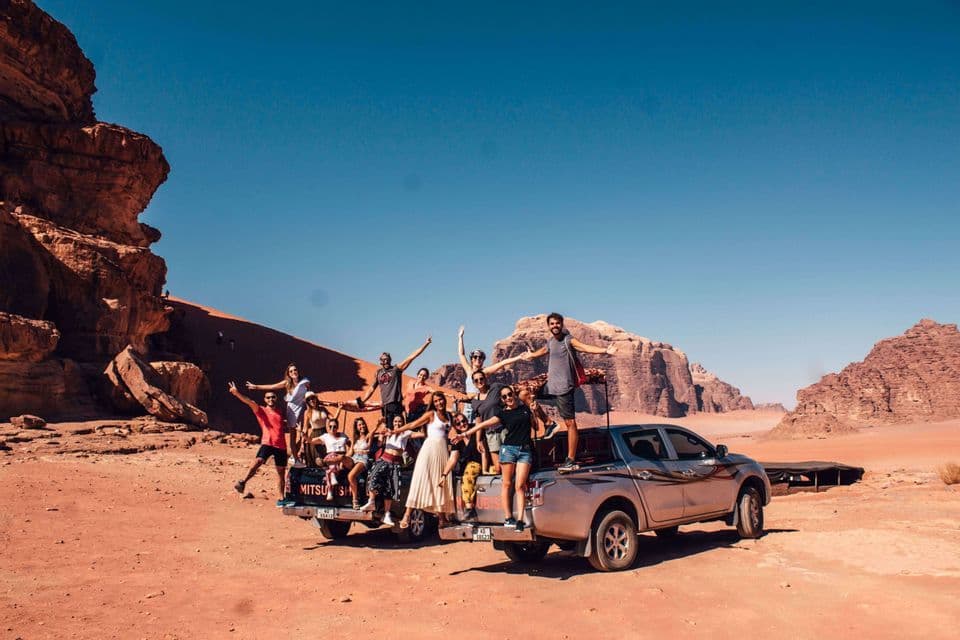 A WeRoad group trip poses for a photo on the back of a pickup truck in a vast desert surrounded by red rock formations.