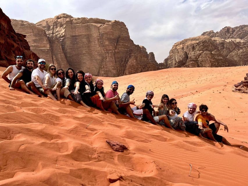 A WeRoad group trip sits in a single file line on a red sand dune, smiling in front of large rock formations.