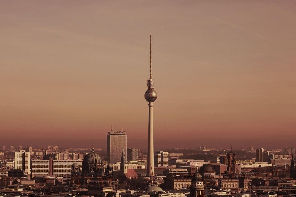 La Torre de Televisión de Berlín se alza sobre el paisaje urbano, que incluye la cúpula de una catedral, todo bañado en la cálida luz de un atardecer brumoso.