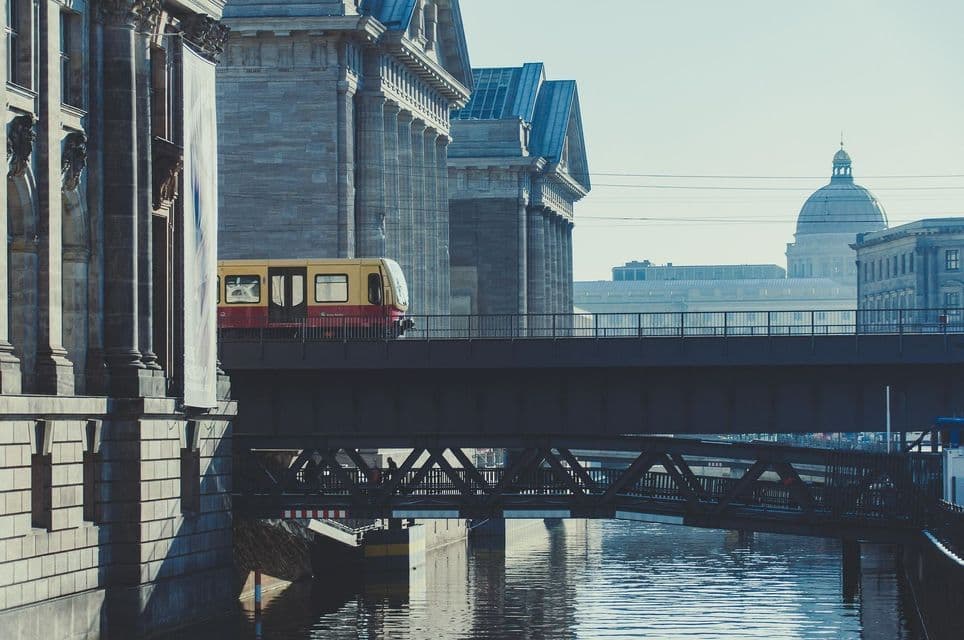 Un tren amarillo y rojo cruza un puente sobre un canal, con grandes edificios de piedra y una estructura abovedada al fondo.