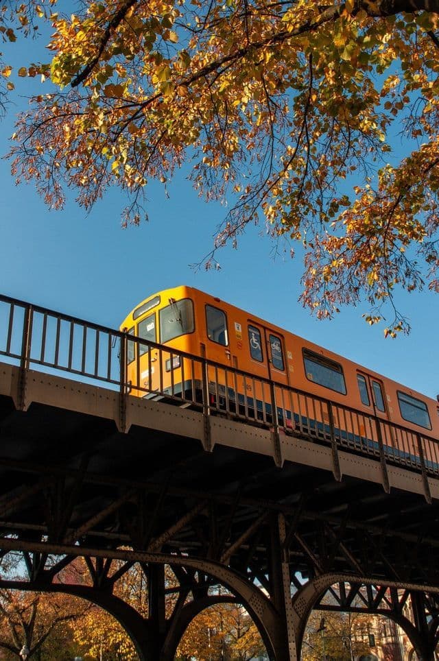 Un tren naranja brillante cruza un puente metálico elevado, enmarcado por hojas amarillas de otoño contra un cielo azul claro.