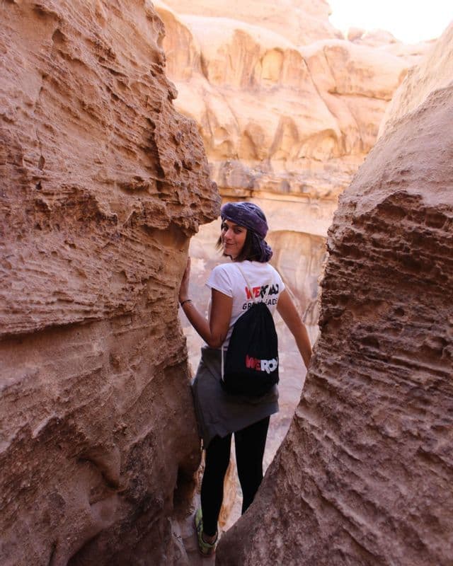 A woman wearing a WeRoad t-shirt and backpack looks back and smiles while walking through a narrow rock canyon.