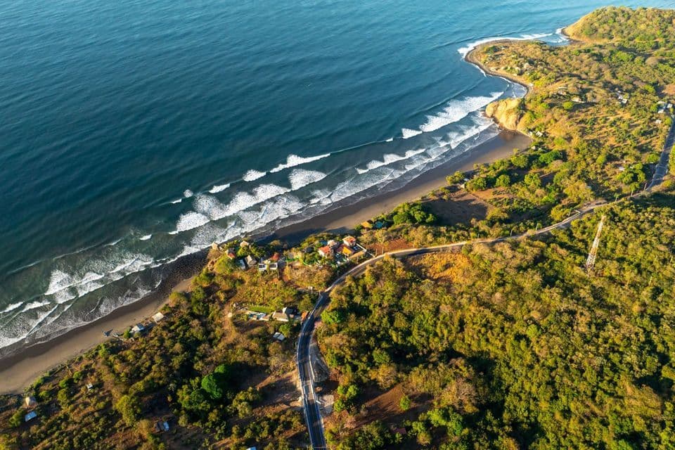 Luftaufnahme einer üppigen, grünen Küste mit einer sich schlängelnden Straße, neben einem Sandstrand, wo weiße Wellen aus dem blauen Meer brechen.