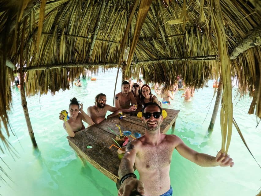 Un gruppo WeRoad in viaggio si scatta un selfie mentre sorseggia un drink a un tavolo parzialmente sommerso in acqua turchese, sotto un tetto di paglia.