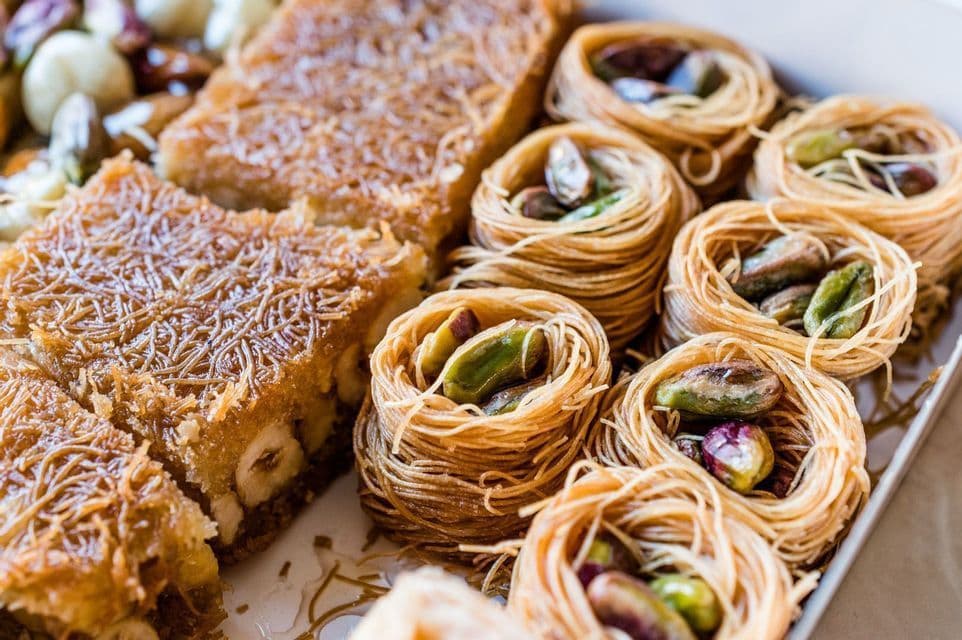 Gros plan sur un assortiment de pâtisseries du Moyen-Orient, comprenant des nids de pâte effilée garnis de pistaches et des baklavas carrés aux noisettes.