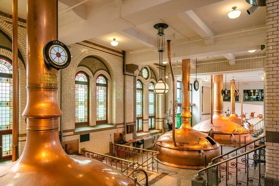 L'intérieur d'une brasserie traditionnelle avec de grandes cuves de brassage en cuivre, des murs carrelés blancs et des vitraux en arc.