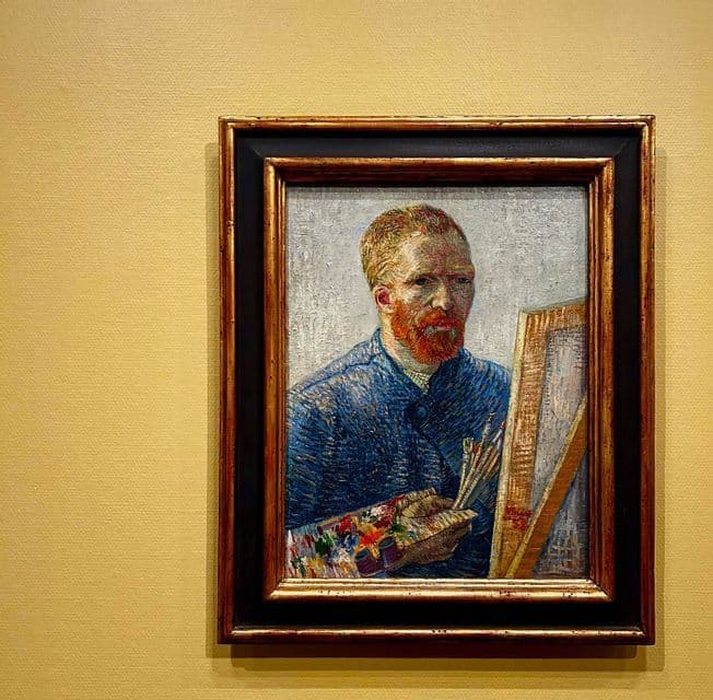 Un tableau encadré d'un autoportrait de Vincent van Gogh, tenant une palette et des pinceaux, est accroché sur un mur jaune.