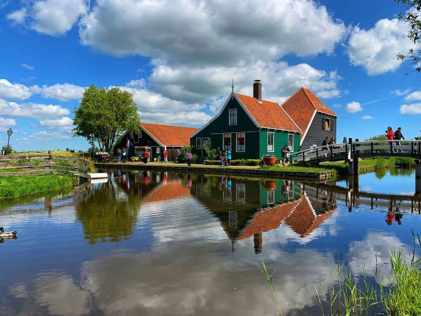 Casas de madera verdes con techos de tejas naranjas se reflejan en las tranquilas aguas de un canal bajo un cielo azul nublado.