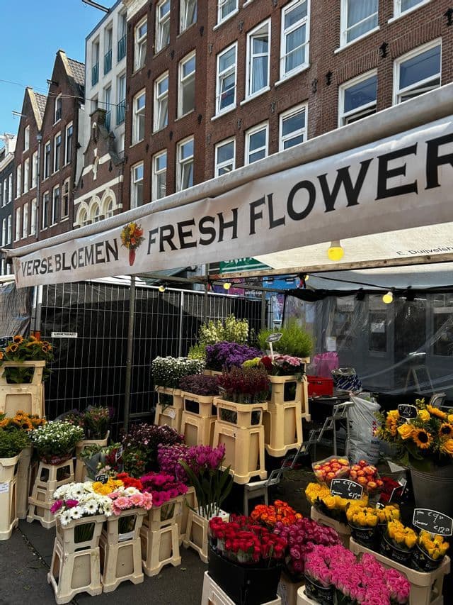 Ein Blumenmarktstand im Freien zeigt verschiedene bunte Blumen in Kisten unter einer Markise mit der Aufschrift "FRISCHE BLUMEN".