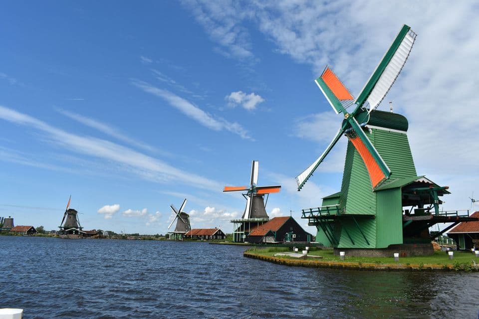 Molinos de viento tradicionales, uno pintado de verde brillante, se alzan en la orilla cubierta de hierba de una vía fluvial bajo un cielo azul.