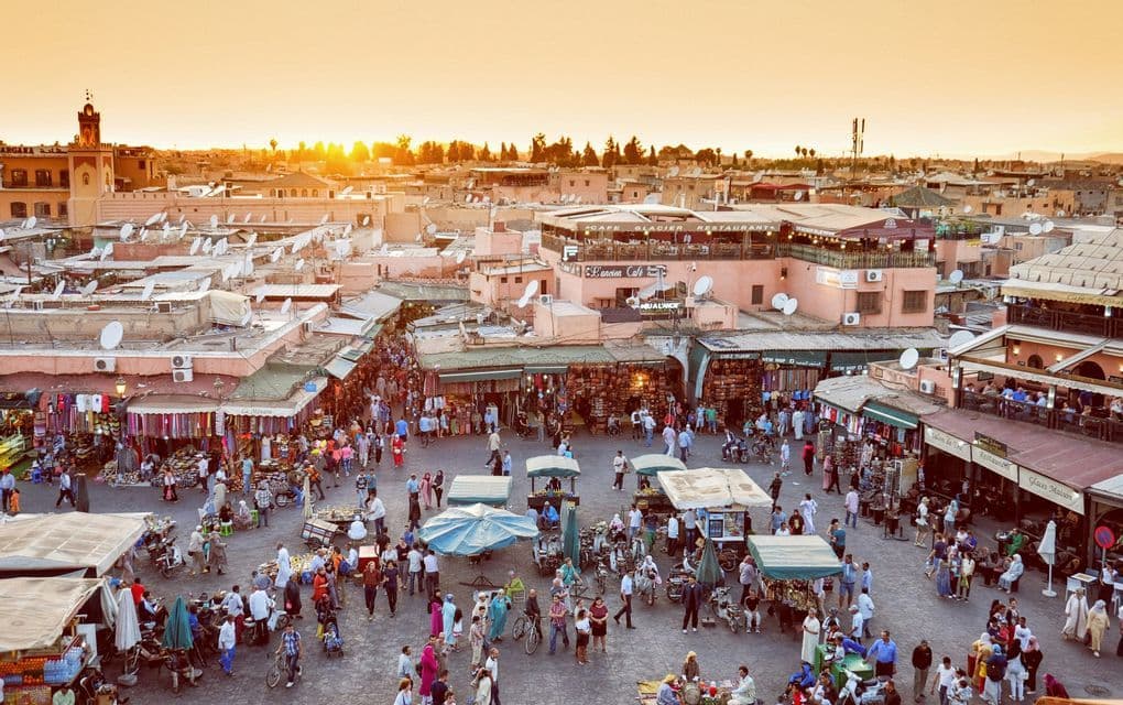 An elevated view of a bustling city square filled with people, market stalls, and shops at sunset.