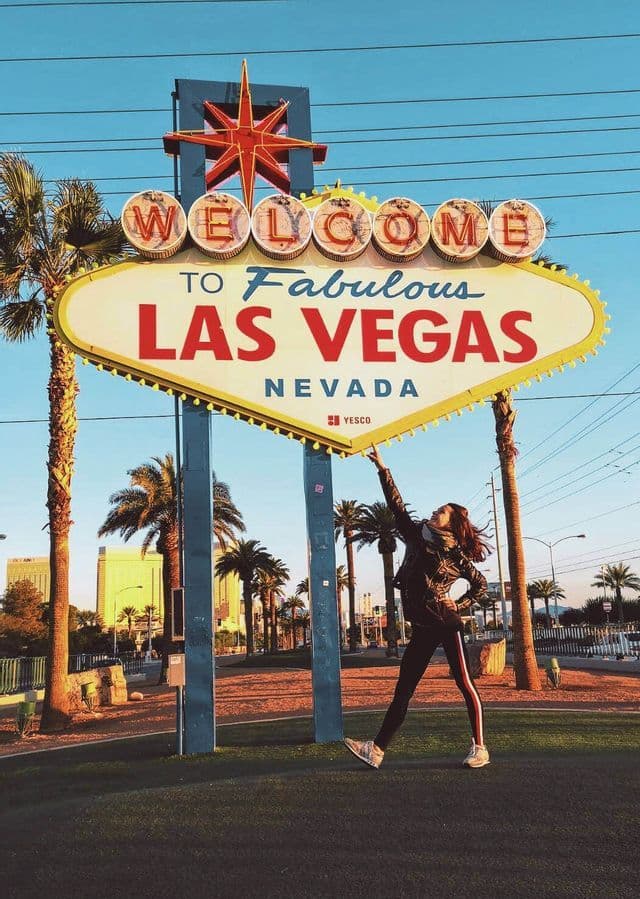 A woman poses with her arm raised, pointing towards the iconic 'Welcome to Fabulous Las Vegas' sign surrounded by palm trees.