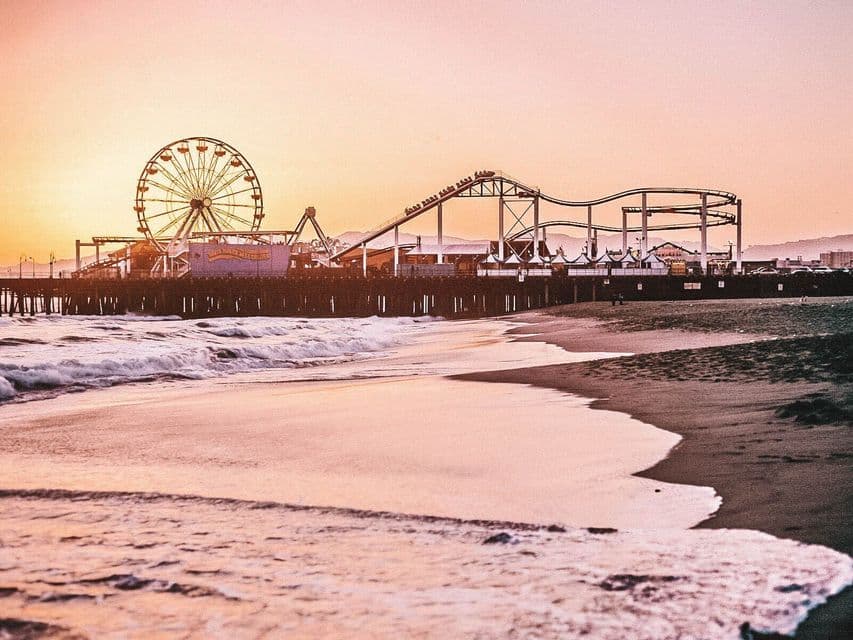 An amusement park with a Ferris wheel and roller coaster on a pier, viewed from a sandy beach at sunset.
