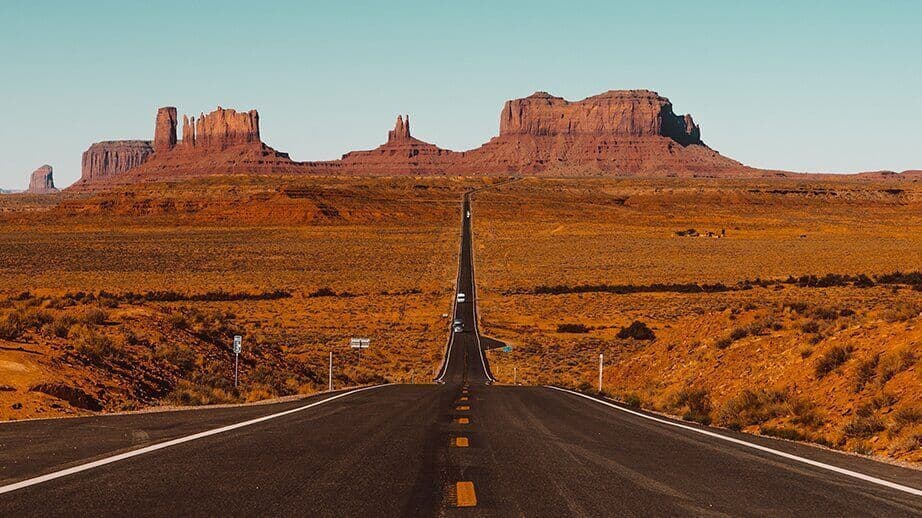 Une longue route rectiligne s'étire à travers un paysage désertique vers d'imposantes buttes de roche rouge sous un ciel bleu pur.