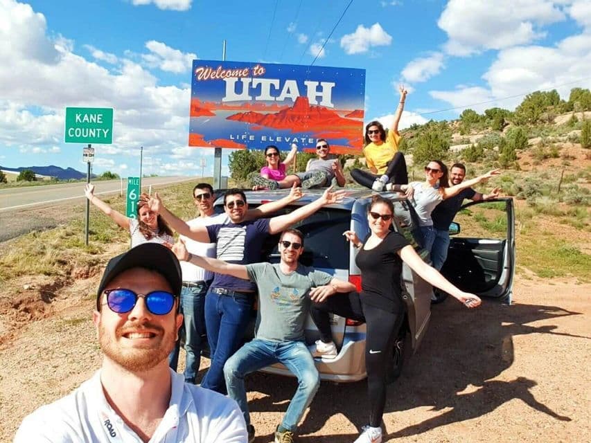 A WeRoad group trip posing for a smiling selfie with their car in front of a 'Welcome to Utah' road sign.