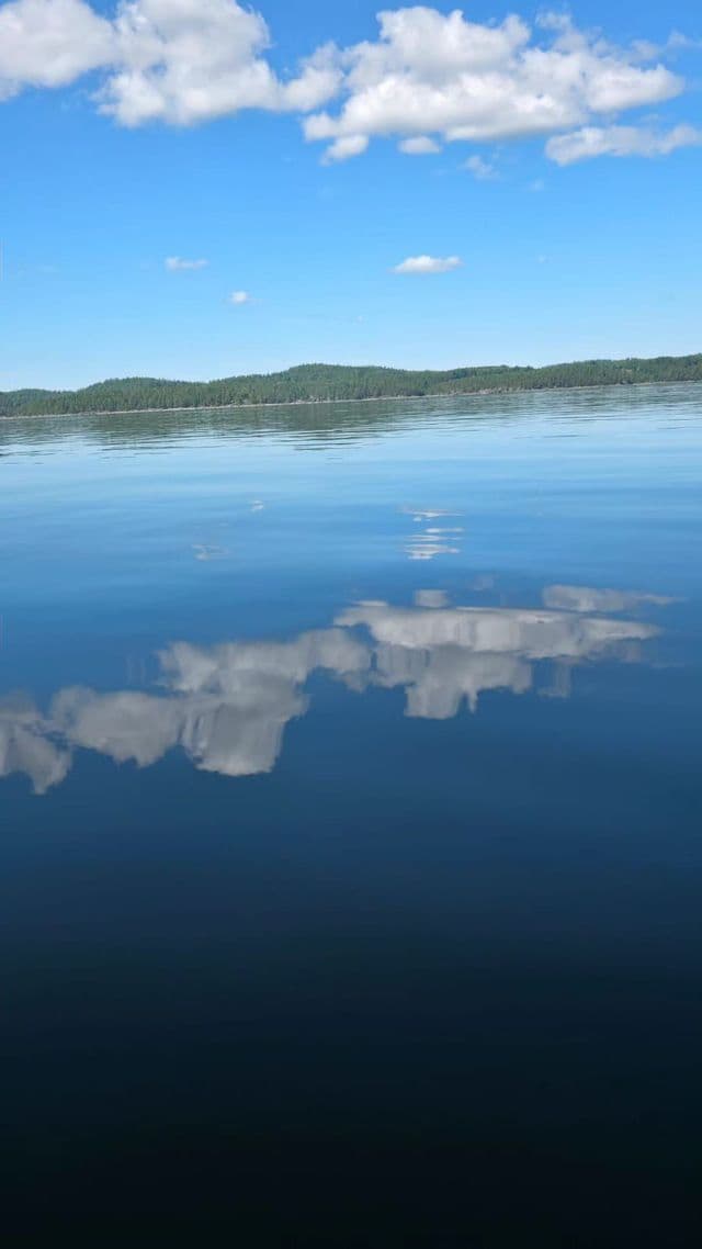 Ein ruhiger See spiegelt perfekt die flauschigen weißen Wolken in einem hellblauen Himmel wider, mit einem bewaldeten Hügel in der Ferne.