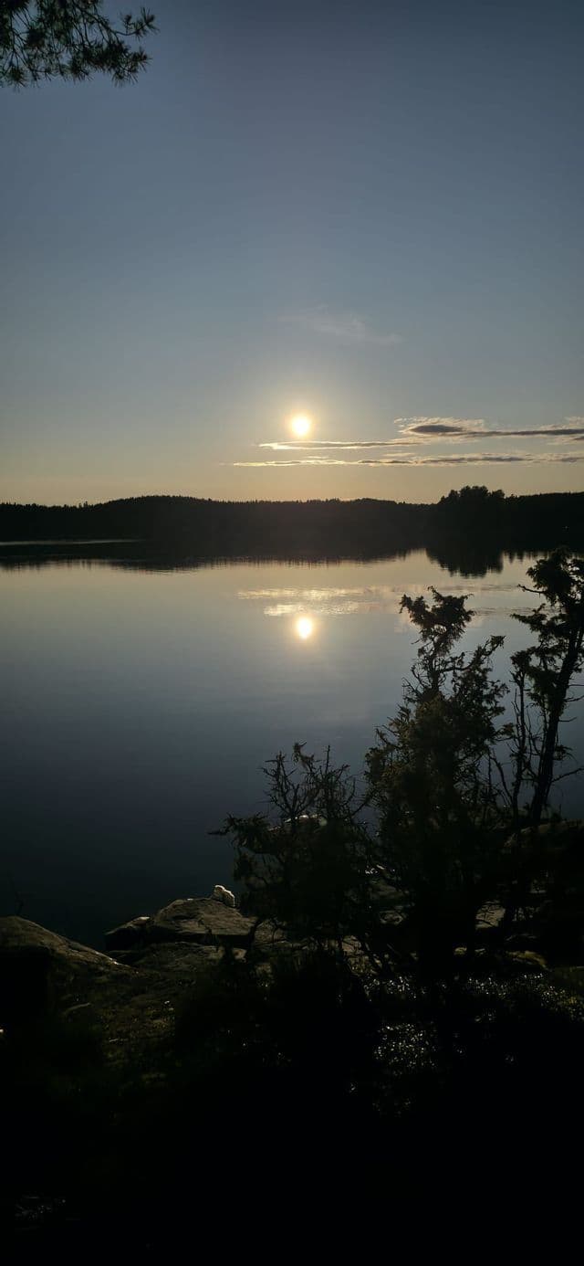 Die Sonne geht über einem ruhigen See unter, ihr Licht spiegelt sich auf dem Wasser vor einem schemenhaften Wald am gegenüberliegenden Ufer.