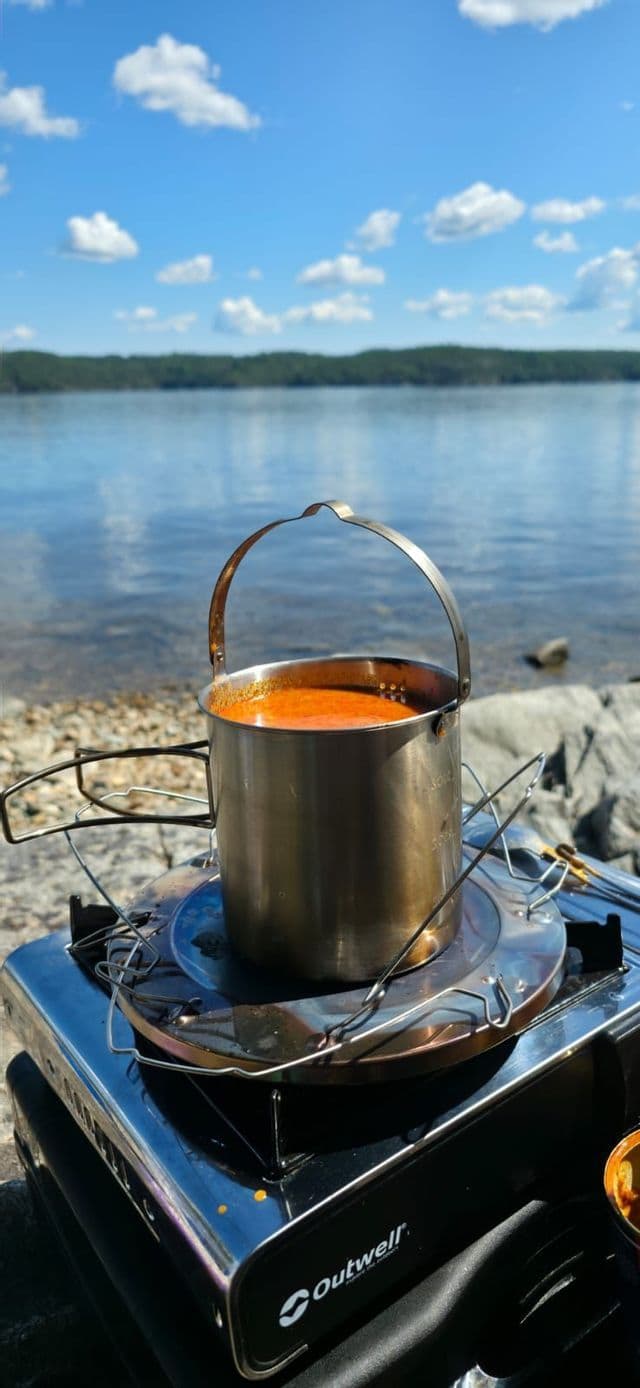 Ein Metalltopf mit orangefarbener Suppe kocht auf einem tragbaren Kocher am felsigen Ufer eines Sees unter blauem Himmel.