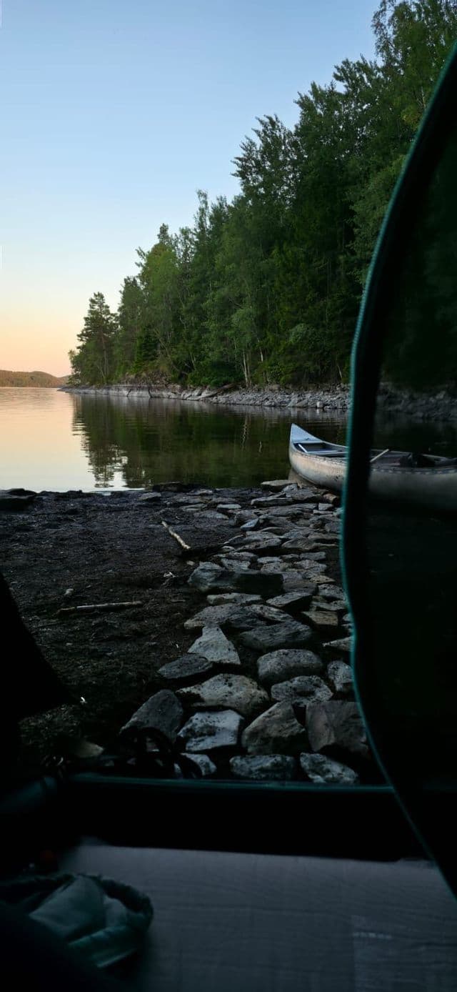 Blick aus einem Zelt auf ein Kanu am felsigen Ufer eines ruhigen, von Wald umgebenen Sees bei Sonnenuntergang.