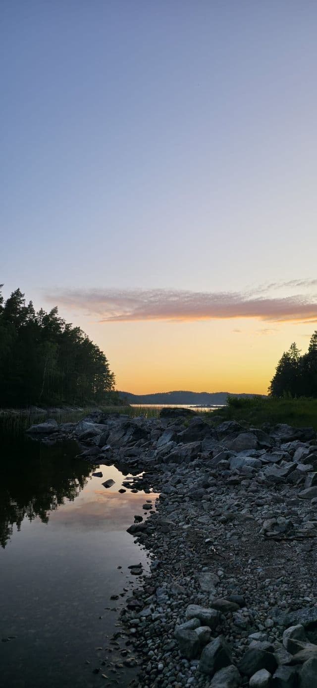 Felsige Uferlinie an einem ruhigen See, in dem sich ein Wald bei farbenprächtigem Sonnenuntergang spiegelt.