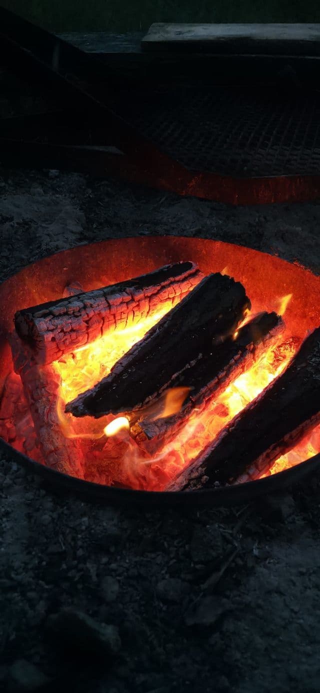 Eine Nahaufnahme von brennenden Holzscheiten mit glühenden orangefarbenen Glutstücken in einer runden Metall-Feuerschale bei Nacht.