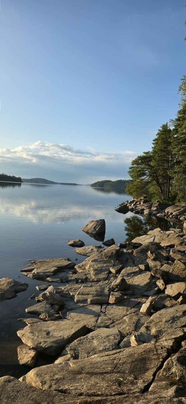 Ein ruhiger See mit felsigem Ufer im Vordergrund, der den blauen Himmel und Wolken spiegelt, gesäumt von Kiefern am Ufer.