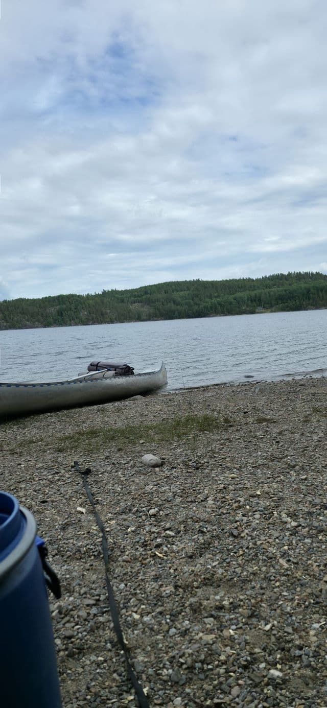 Ein Kanu mit Ausrüstung liegt an einem kiesigen Seeufer, mit einem bewaldeten Hügel auf der anderen Wasserseite unter bewölktem Himmel.