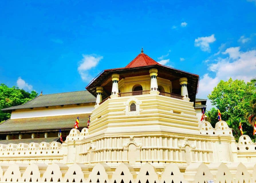 Ein mehrstufiger weißer Tempel mit einem achteckigen Turm und rotem Dach steht hinter einer dekorativen Mauer unter blauem Himmel.