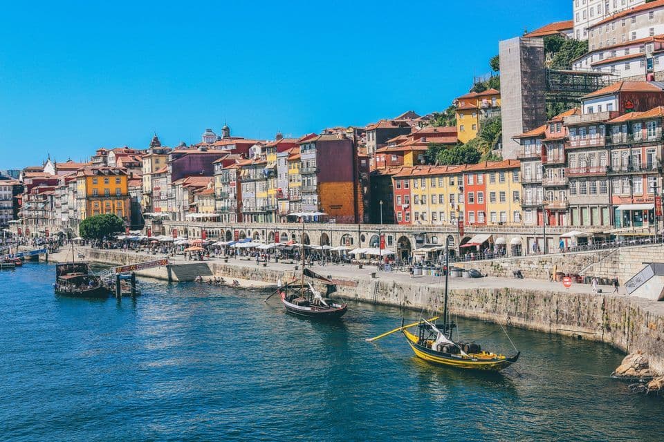 Des bateaux traditionnels flottent sur une rivière le long d'un front de mer animé, bordé de bâtiments colorés et historiques, sous un ciel bleu clair.