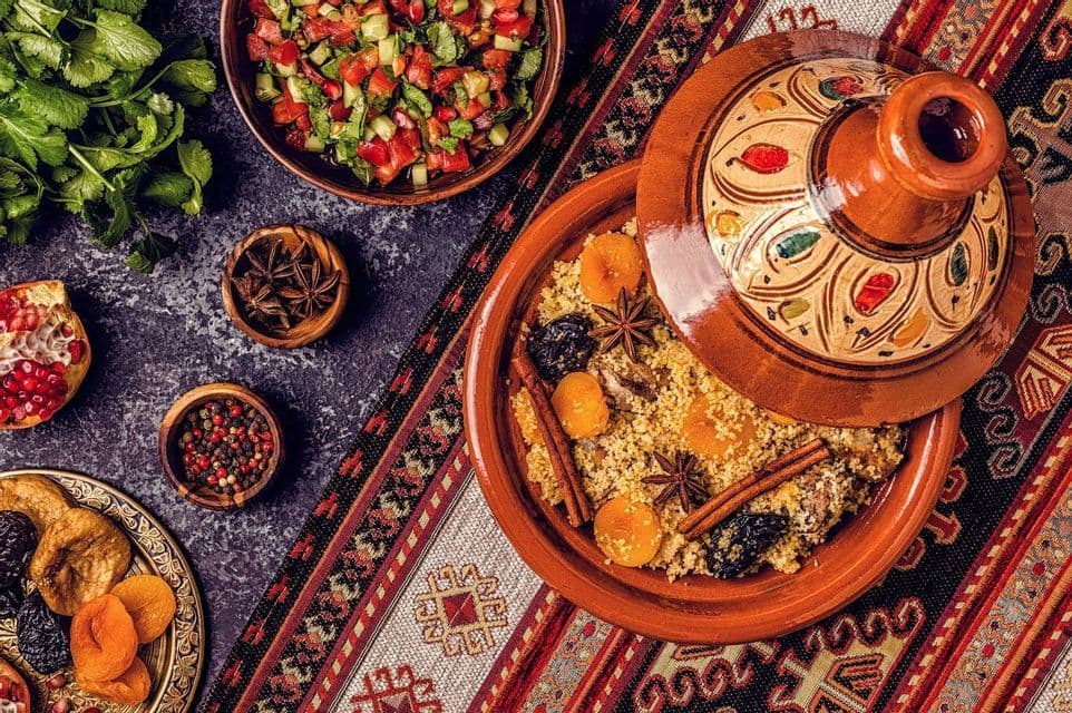 A Moroccan tagine filled with couscous, dried fruit, and spices rests on a patterned cloth next to a bowl of fresh salad.
