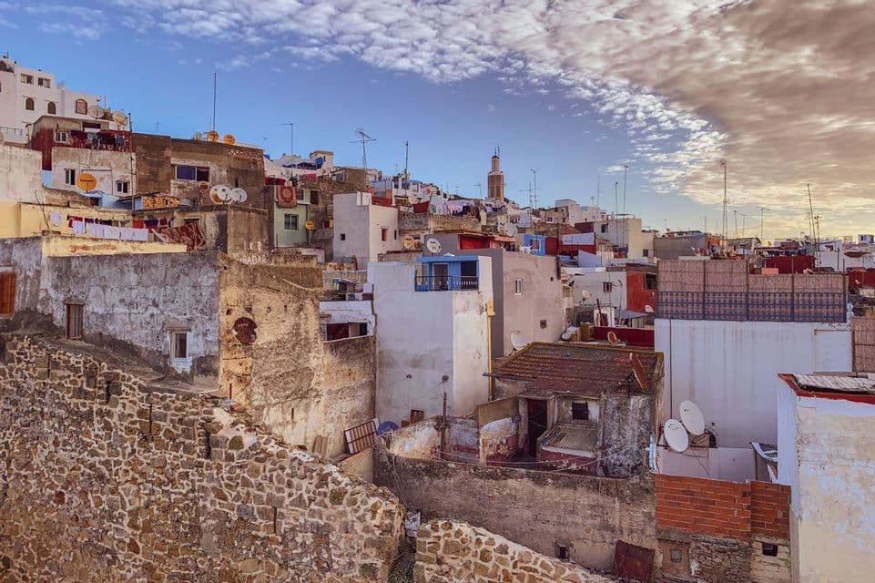 A dense cityscape of weathered buildings on a hillside, with a distant minaret under a partly cloudy sky.