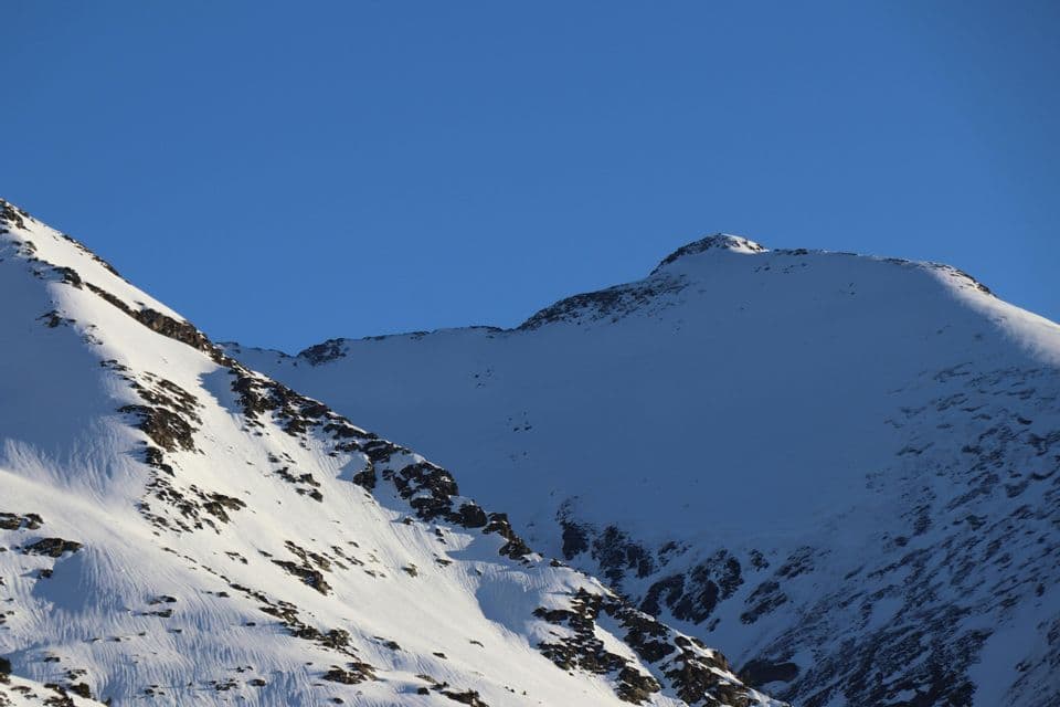 Picos montañosos nevados y soleados con formaciones rocosas oscuras resaltan contra un cielo azul claro