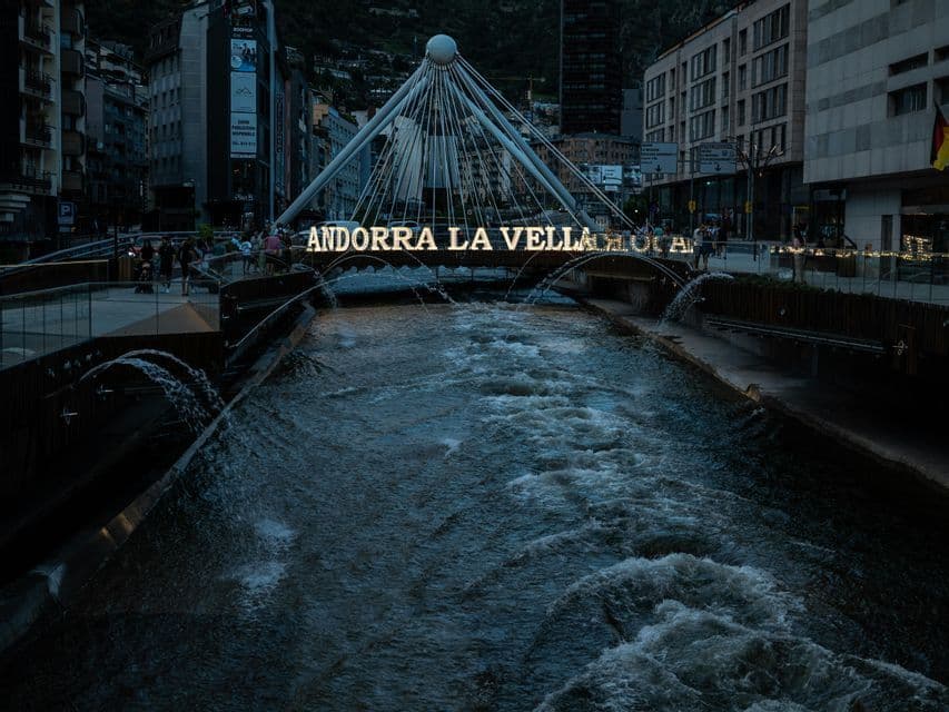 Un letrero iluminado de 'ANDORRA LA VELLA' en un puente blanco sobre un río caudaloso con fuentes al atardecer.