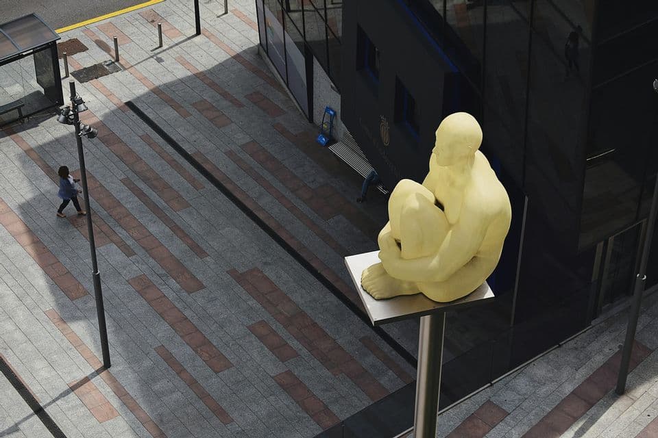 Vista en picada de una gran escultura amarilla de una figura sentada que domina una calle urbana pavimentada por donde camina una persona.