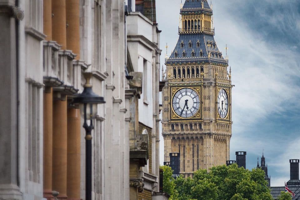 La torre del reloj Big Ben se ve desde una calle, parcialmente enmarcada por edificios de piedra y copas de árboles bajo un cielo nublado.