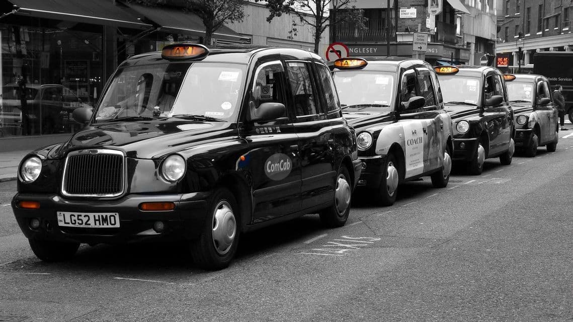 Una fila de taxis negros estilo londinense aparcados en una calle de la ciudad, en una foto en blanco y negro con coloración naranja selectiva en los letreros de los taxis.
