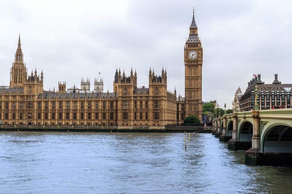 El Palacio de Westminster y la torre del reloj Big Ben vistos desde el otro lado del río Támesis, con un puente de piedra a la derecha, bajo un cielo nublado.