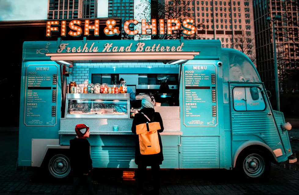 Un adulto y un niño se encuentran en el mostrador de un food truck vintage, azul claro, con un letrero de 'Fish & Chips' en un entorno urbano.