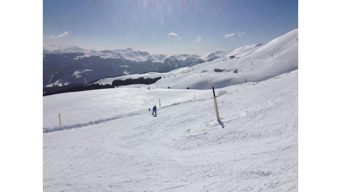 Uno sciatore in giacca blu scende un ampio pendio montano innevato sotto un cielo soleggiato.