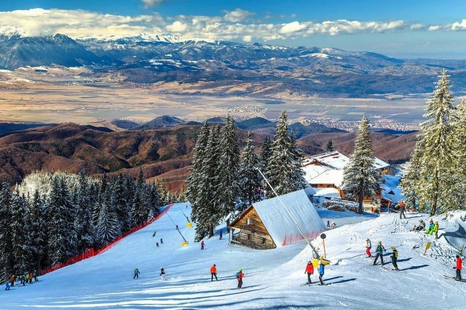 Una vista aerea di persone che sciano su una pista in una località montana, con chalet innevati che si affacciano su un'ampia valle e montagne distanti.