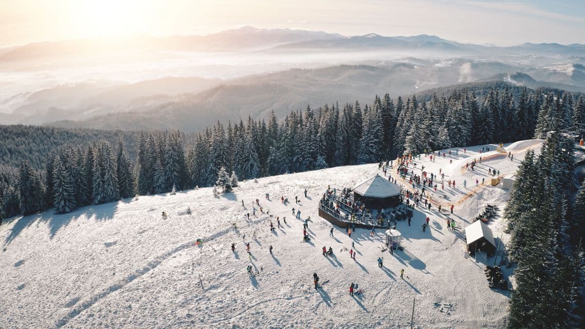 Luftaufnahme von vielen Menschen, die an einer sonnigen, schneebedeckten Bergflanke neben einer Hütte und einem dichten Föhrenwald Ski fahren.