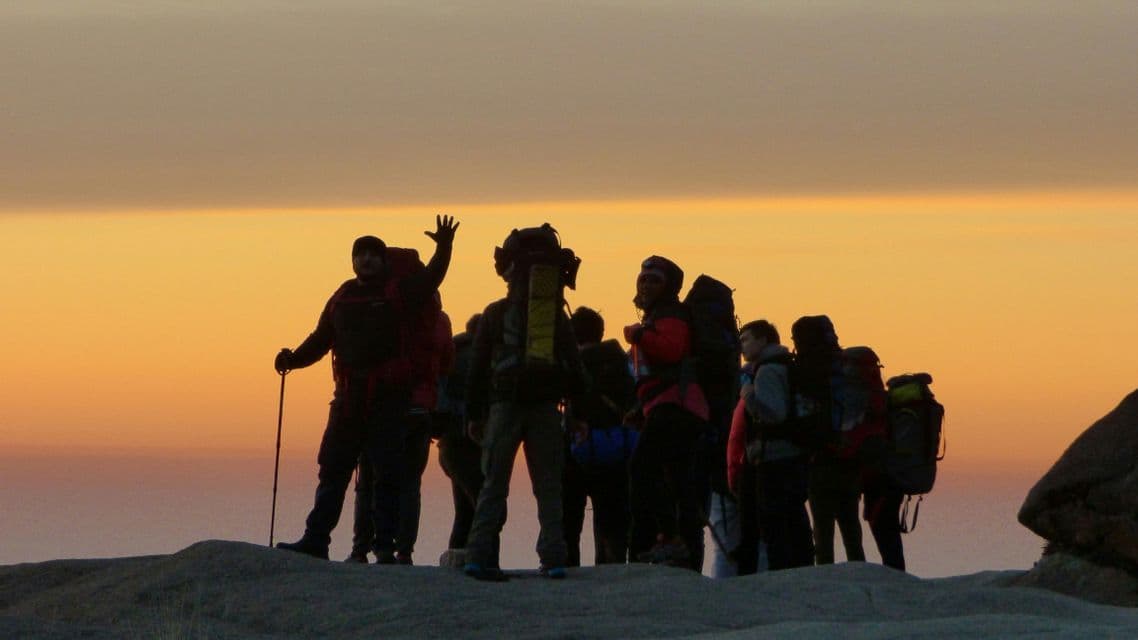 Eine WeRoad-Gruppe von Wanderern mit Rucksäcken ist auf einem Hügel gegen einen goldenen Himmel bei Sonnenuntergang silhouettiert.