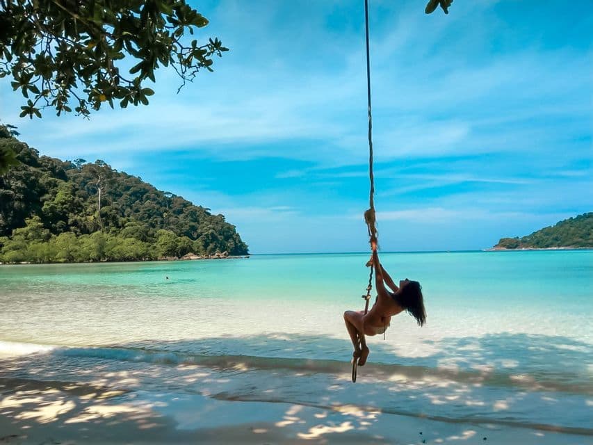 A woman in a bikini on a rope swing over a tropical beach with white sand and clear turquoise water.