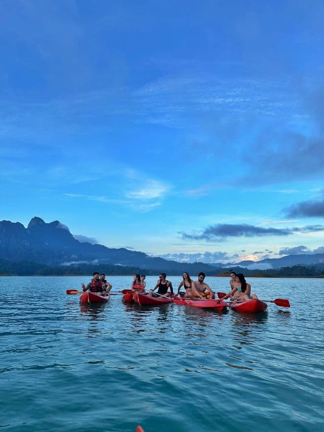 A WeRoad group trip kayaking on a lake in red boats, with misty mountains in the background under a blue sky.