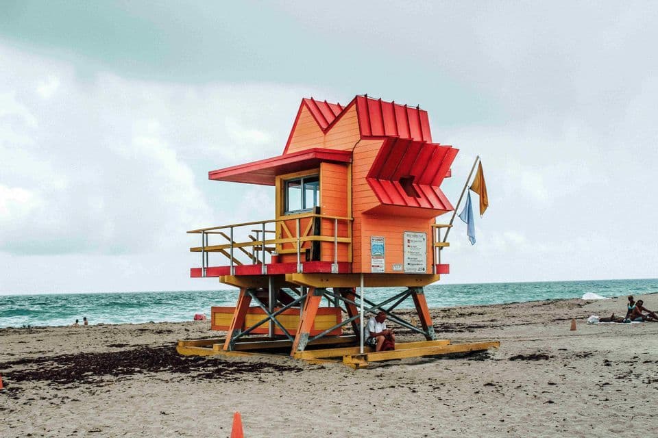 Una colorida torre de salvavidas naranja y roja se alza en una playa de arena junto al océano bajo un cielo nublado.